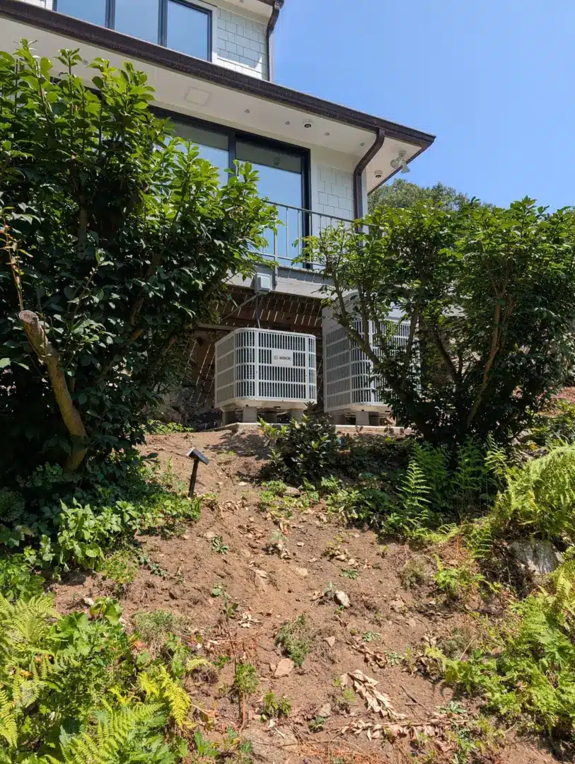 Two Bosch heat pump outdoor units installed below a modern home on a wooded hillside in Greenwich, CT by Celco